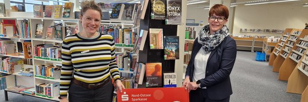 Zwei Frauen halten einen großen symbolischen Scheck hoch mit dem Betrag 500 Euro. Im Hintergrund sind viele Bücherregale mit Büchern bestückt