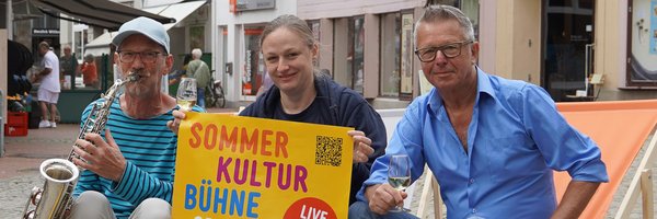 Drei Personen auf Liegestühlen auf dem Kornmarkt mit dem Plakat der Veranstaltung