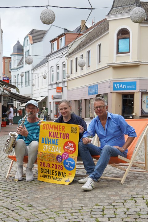 Drei Personen auf Liegestühlen auf dem Kornmarkt mit dem Plakat der Veranstaltung