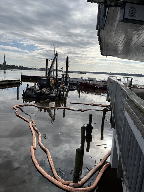 Containerschiff mit Bagger. Im Wasser sind Ölsperren und kleinteilige Wrackreste zu sehen.
