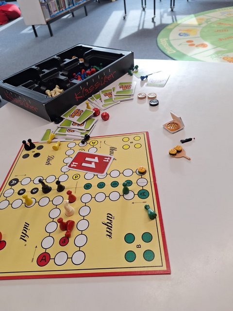 Mensch-ärgere-dich-nicht-Spiel mit Spielfiguren, Skip-Bo- und 11er-raus-Karten, Würfel und Miniaturpizzen auf einem Tisch