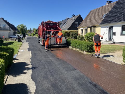 Asphaltarbeiten im Heisterweg, eine geteerte Straße und Bauarbeiter auf einem Fahrzeug von hinten