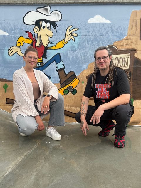 Streetworker*innen Schleswig - Marie-Louise Finger und Tobias H. Müller vor einer Sprayerwand, auf der Lucky Luke zu sehen ist.