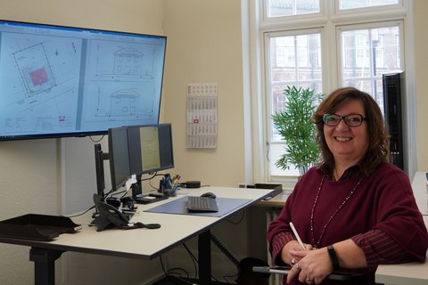 Anja Friedrich stehend an ihrem Arbeitsplatz, vor ihr der höhenverstellbare Schreibtisch und zwei Bildschirme