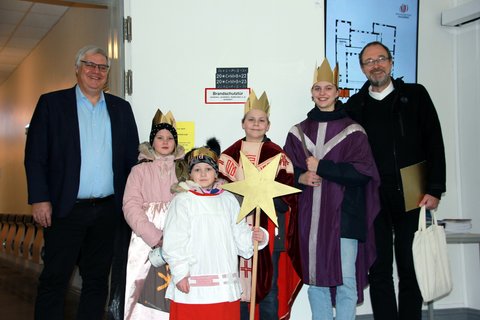 Das Bild zeigt die Sternsinger mit Pastor Winter und dem Bürgermeister der Stadt Schleswig Stephan Dose. Im Hintergrund ist das an eine Tür geklebte Kreidezeichen "20*C+M+B+24" zu erkennen, mit dem der Segen Gottes in das Schleswiger Rathaus gebracht wird.
