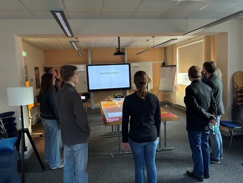 Menschen stehen in einem Raum mit dem Gesicht zu einem Bildschirm, worauf "Das Spiel beginnt" zu lesen ist