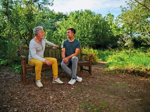 Zwei Männer sitzen auf einer Bank im Grünen und schauen sich an.