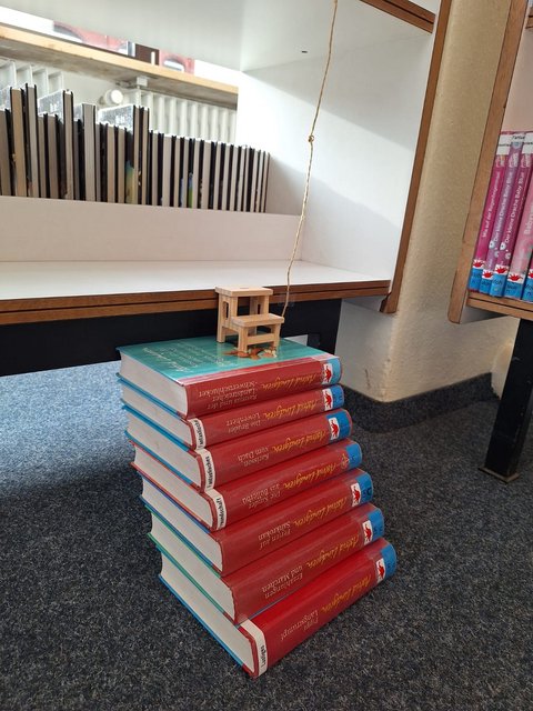 Sieben Bücher übereinandergestapelt, ganz oben steht ein Miniaturstuhl vor einem Bücherregal