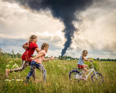 Das Bild zeigt drei Kinder. Rechts fährt ein Mädchen auf einem Fahrrad auf einer Wiese und links laufen zwei Mädchen hinterher. Im Hintergrund ist schwarzer am Horizont aufsteigender Rauch zu sehen. Es ist das UNICEF-Foto des Jahres.