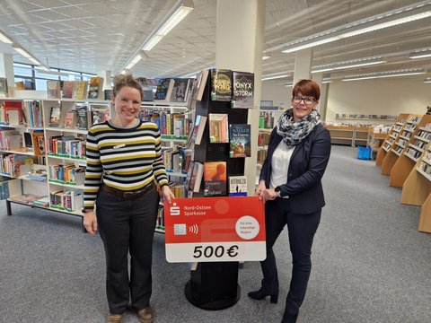 Zwei Frauen halten einen großen symbolischen Scheck hoch mit dem Betrag 500 Euro. Im Hintergrund sind viele Bücherregale mit Büchern bestückt
