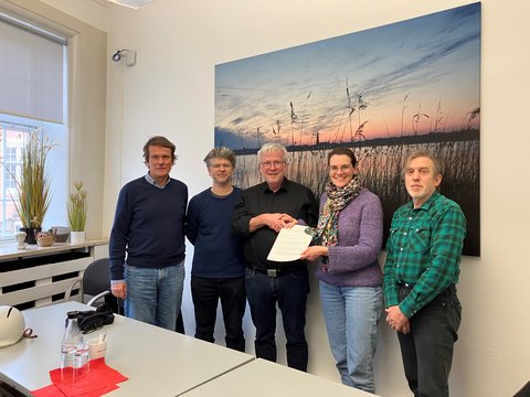 Vertragsunterzeichnung Energiegemeinschaft Schlei und Stadt Schleswig: Michael Struck, Robert Wünsch, Bernward Völmicke (Stadt Schleswig), Cornelia Sperling, Peter Kieseritzky stehen vor einem großen Bild mit Abendlandschaft