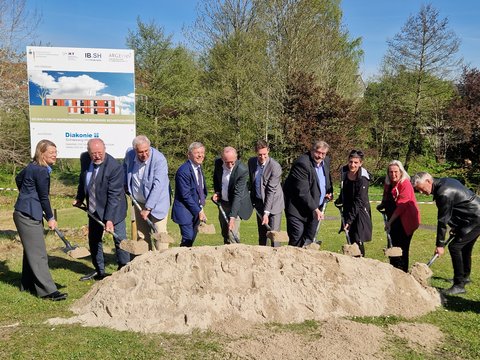 Spatenstich für ein zukunftsweisendes Bauprojekt im Ansgarweg 4–5: Vertreter*innen der Diakonie Stiftung Schleswig-Holstein, des Landes S-H, des Kreises Schleswig-Flensburg und der Stadt Schleswig. Alle Personen haben einen Spaten in der Hand und stechen in den Sandhaufen vor ihnen. Im Hintergrund Bauschild und Bäume
