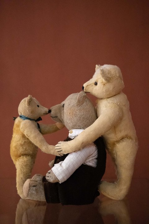 Links ein kleinerer Teddybär, mit dem Körper nach rechts, einem größeren, sitzenden Teddybären zugewandt. Ganz Rechts ein weiterer größerer Teddybär, der den sitzenden an den Schultern hält. 