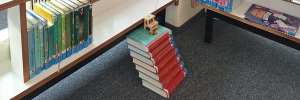 Sieben Bücher übereinandergestapelt, ganz oben steht ein Miniaturstuhl vor einem Bücherregal mit sehr vielen Büchern