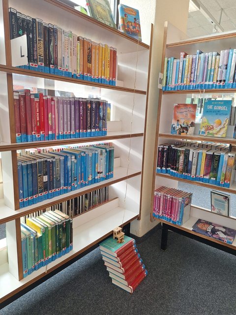 Sieben Bücher übereinandergestapelt, ganz oben steht ein Miniaturstuhl vor einem Bücherregal mit sehr vielen Büchern