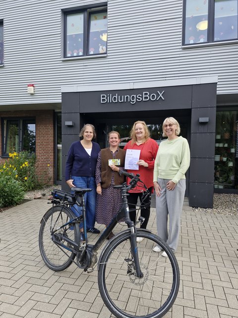 Übergabe Preisgeld Kita BildungsBox: B. Hindrichsen, F. Großkopf und S. Struve von der Kita BildungsBox sowie N. Wiemeier (Stadt Schleswig) vor dem Gebäude der Kita BildungsBox mit einem Fahrrad im Vordergrund