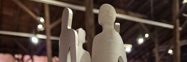 Ein silberner Pokal in Form von menschlichen Silhouetten auf einem runden Podest. 