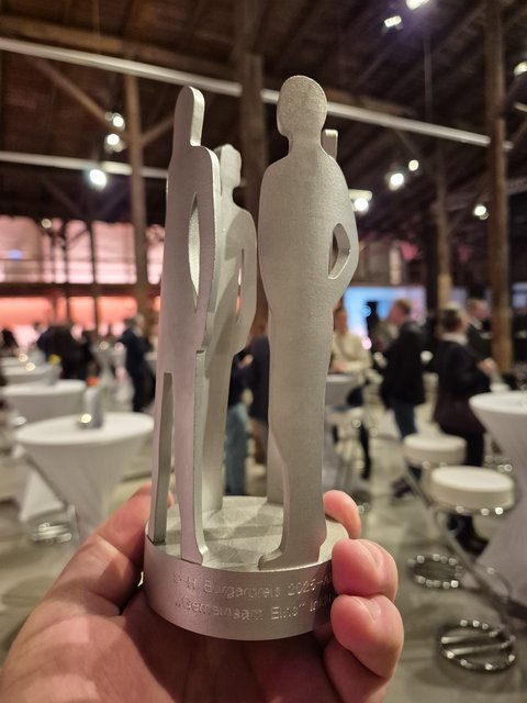 Ein silberner Pokal in Form von menschlichen Silhouetten auf einem runden Podest.