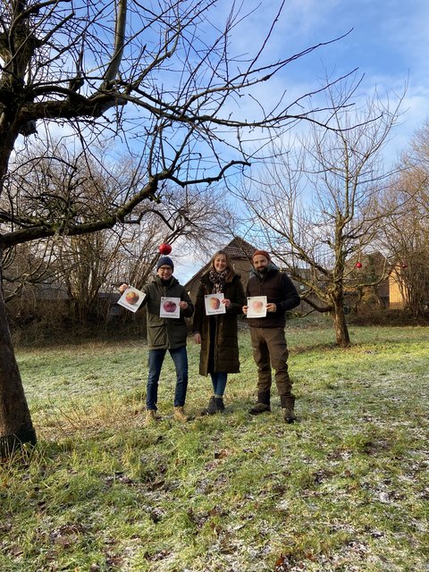 Jens Bagehorn-Delor, (Stadt Schleswig) Maren Thiesen (Naturpark Schlei) und Miklas Staiger (Obstbaumpfleger und Streuobstpädagoge) stehen vor einem Obstbau und halten Schilder mit Äpfeln hoch