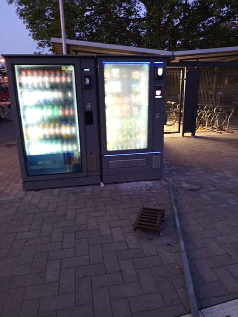 Beschädigter Snackautomat mit gesprungener Scheibe, davor Eisenstange und Gullydeckel auf dem Boden
