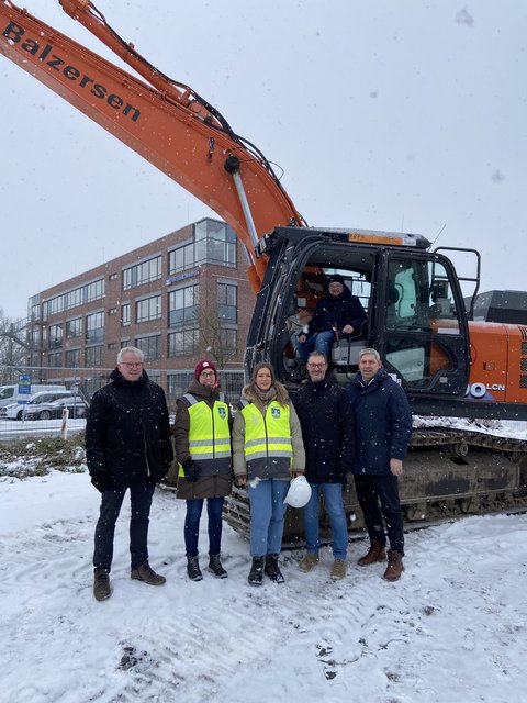 Abbrucharbeiten ZOB-Gebäude 16.02.2026 - Jonas Kähler, Bernward Völmicke, Rowena Sandmeier und Josie Schäfer (Stadt Schleswig), Christian Liesegang (IGL) und Helge Schütze (Stadtmarketing) vor dem Bagger auf dem ZOB-Gelände