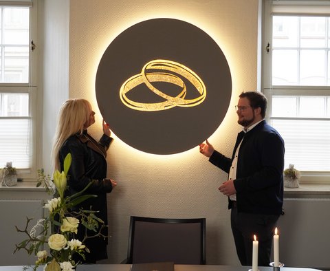 Künstlerin Kathrin Geller und Bürgermeister Jonas Kähler vor dem beleuchteten Kunstobjekt im Trauzimmer, das zwei goldene Ringe zeigt.