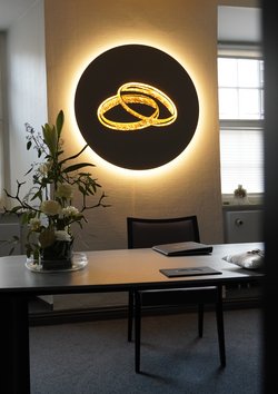 Tisch mit Blumengesteck und der Traumappe darauf. Dahinter steht ein Stuhl und an der Wand ist ein beleuchtetes Kunstwerk mit zwei Ringen in Gold zu sehen.