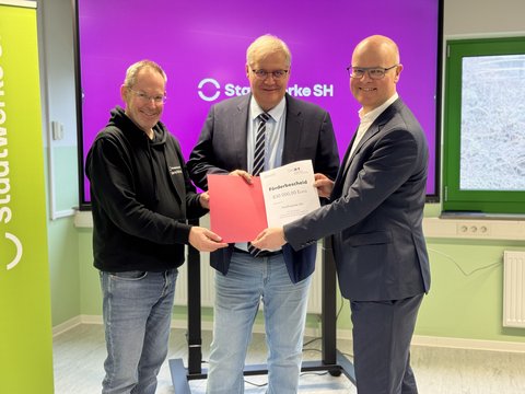 Übergabe Fördermittelbescheid: Bernd Reichelt (Stadtwerke SH), Bürgermeister Stephan Dose und Umweltminister des Landes Schleswig-Holstein, Tobias Goldschmidt halten den Fördermittelbescheid in die Kamera
