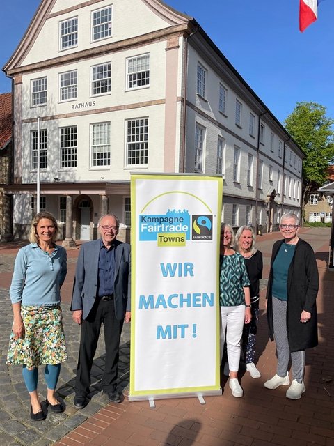 Das Foto zeigt aus dem Steuerungsgruppen-Team Sina Clorius (Ratsversammlung), Matthias Hertel und Ute Ulrich (Verein zur Förderung des fairen Handels Schleswig e. V.)  Maren Korban und Inke Asmussen (beide Ratsversammlung) mit einem Aufsteller "Wir machen mit" vor dem Schleswiger Rathaus