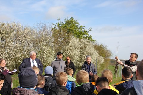 Das Bild zeigt den Bürgermeister der Stadt Schleswig Stephan Dose, Fabian Bellinghausen, Jens Bagehorn-Delor und zwei Grundschulklassen. Gemeinsam schauen sie auf den Bezirksförster Stefan Guntermann, der einen kleinen Baum in der Hand hält.