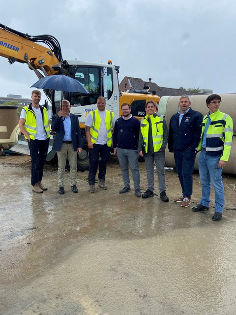 Thore Stamm (Johannssen Straßen-, Hoch- & Tiefbau GmbH), Stefan Wesemann (IHK), Martin Gast (Fa. Johannssen), Jonas Kähler (Stadt Schleswig), Marco Armgort (Masuch + Olbrisch Ingenieurgesellschaft für das Bauwesen mbH), Helge Schütze (Stadtmarketing) und Arne Clemens (Schleswiger Stadtwerke Abwasserentsorgung - Eigenbetreib der Stadt Schleswig) auf der Baustelle am Schwarzen Weg. Im Hintergrund ein Bagger.