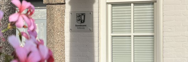 Auf dem Bild ist der Eingang des Standesamtes Schleswig mit dem Schild "Standesamt Schleswig" samt Wappen zu sehen. Im Vordergrund im Anschnitt ist eine Säule und rosa Blüten.