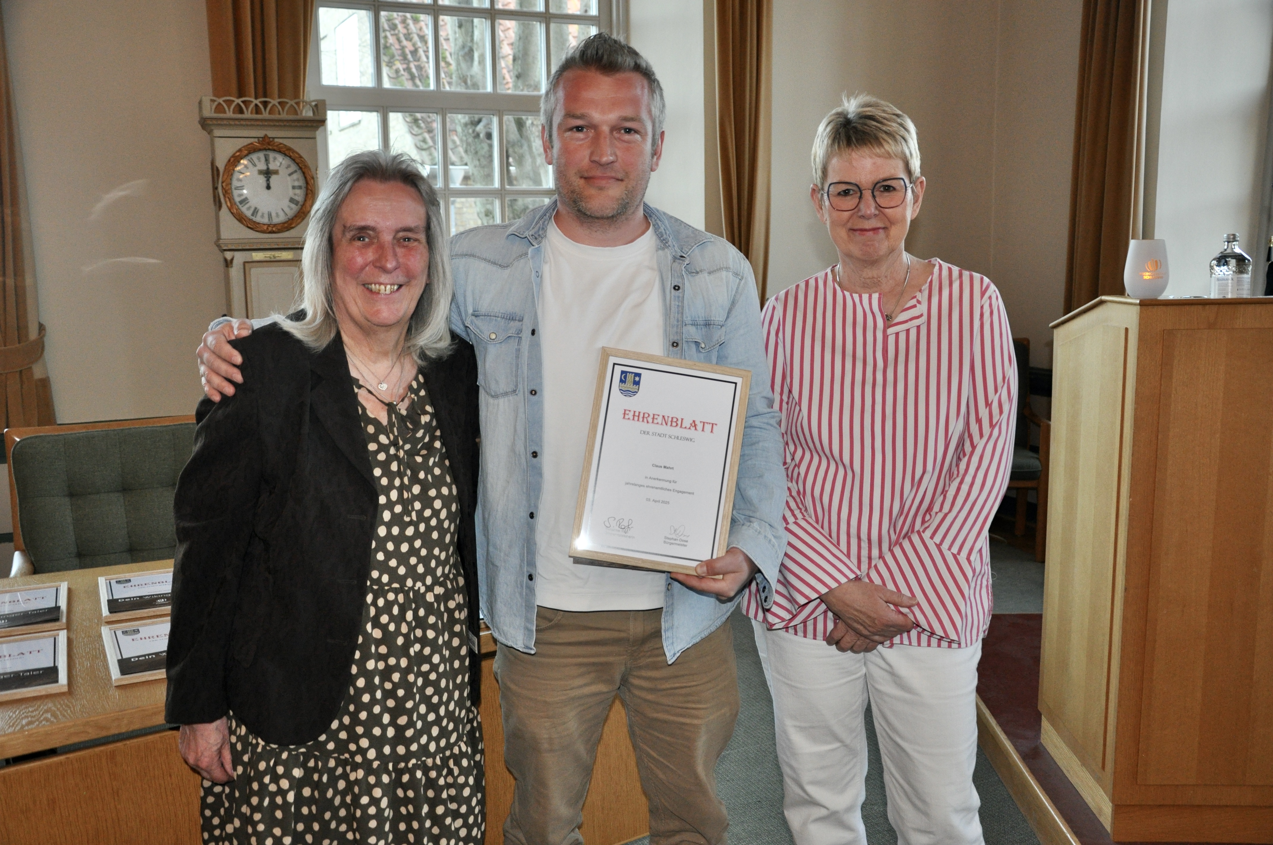 Maren Korban (Vorsitzende des Sozial-, Kultur- und Sportausschusses), Claus Marth (Ehrung verdienter Bürger) und Bürgervorsteherin Susanne Roß stehen im Ständesaal des Rathauses und zeigen die Urkunde