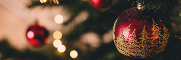 An einem mit einer Lichterkette und Christbaumkugeln geschmückten Baum hängt auf der rechten Seite eine rote Christbaumkugel mit weißen Umrissen von Tannen. Auf dieser liegt der Fokus des Bildes.